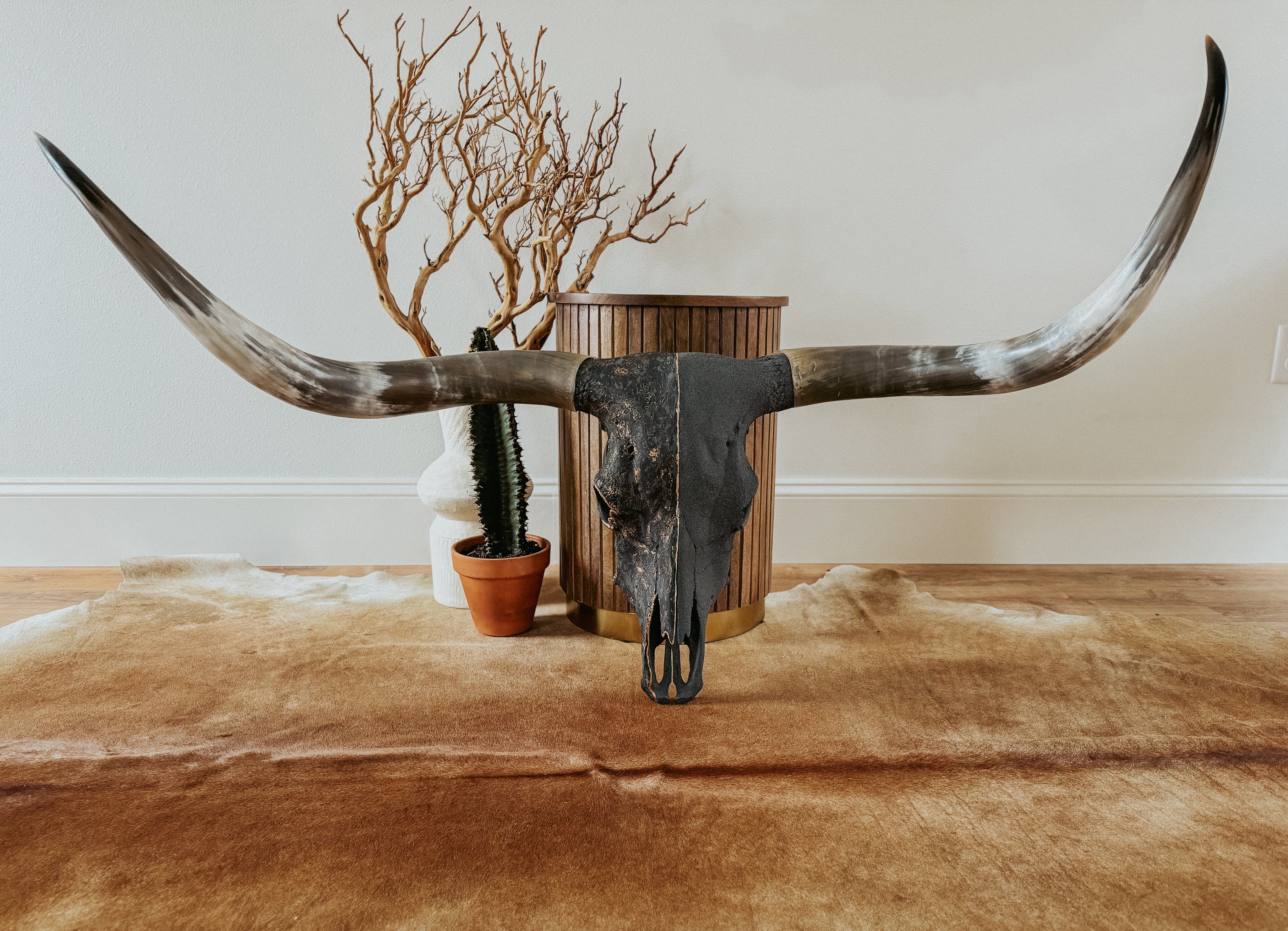 A wall-mounted longhorn skull with asymmetrical horns, featuring a charcoal stone base with heavy black texture and gold glaze, displayed above a cowhide rug.