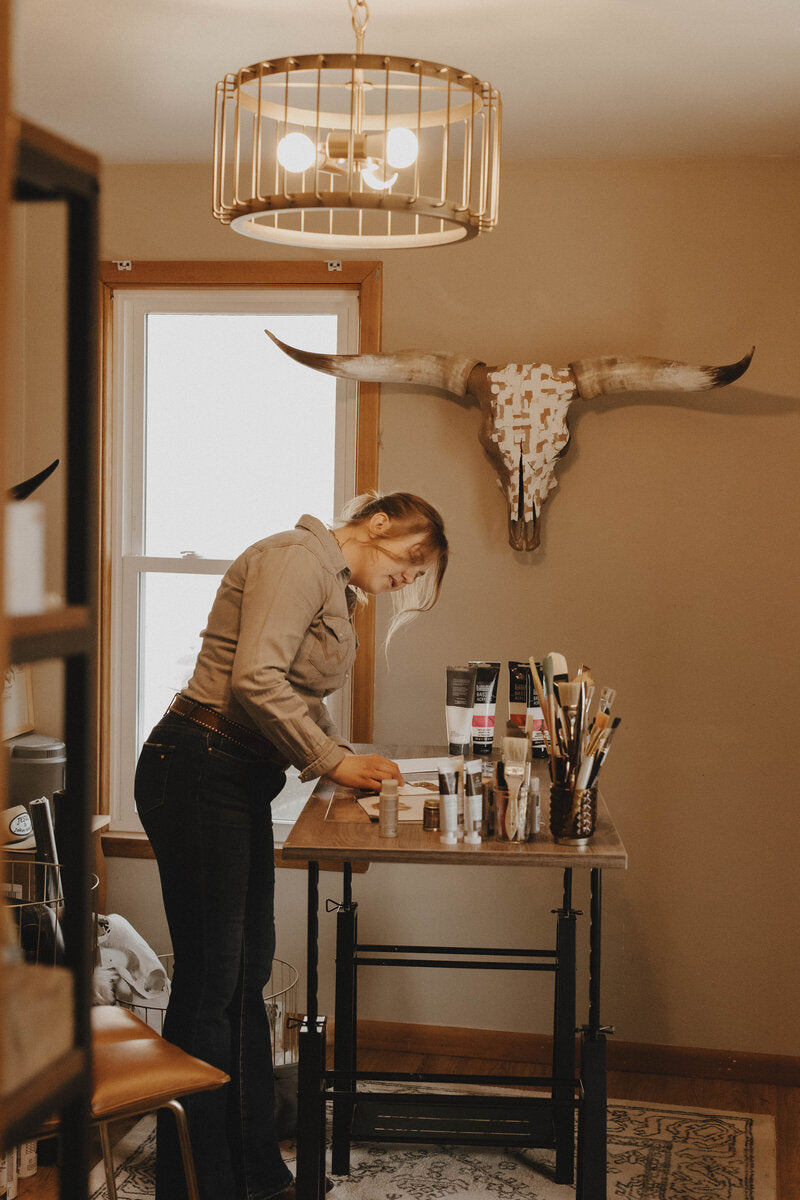 Wanderlust skulls owner designing custom painted animal skulls table with various items in a room with a mounted longhorn skull on the wall.