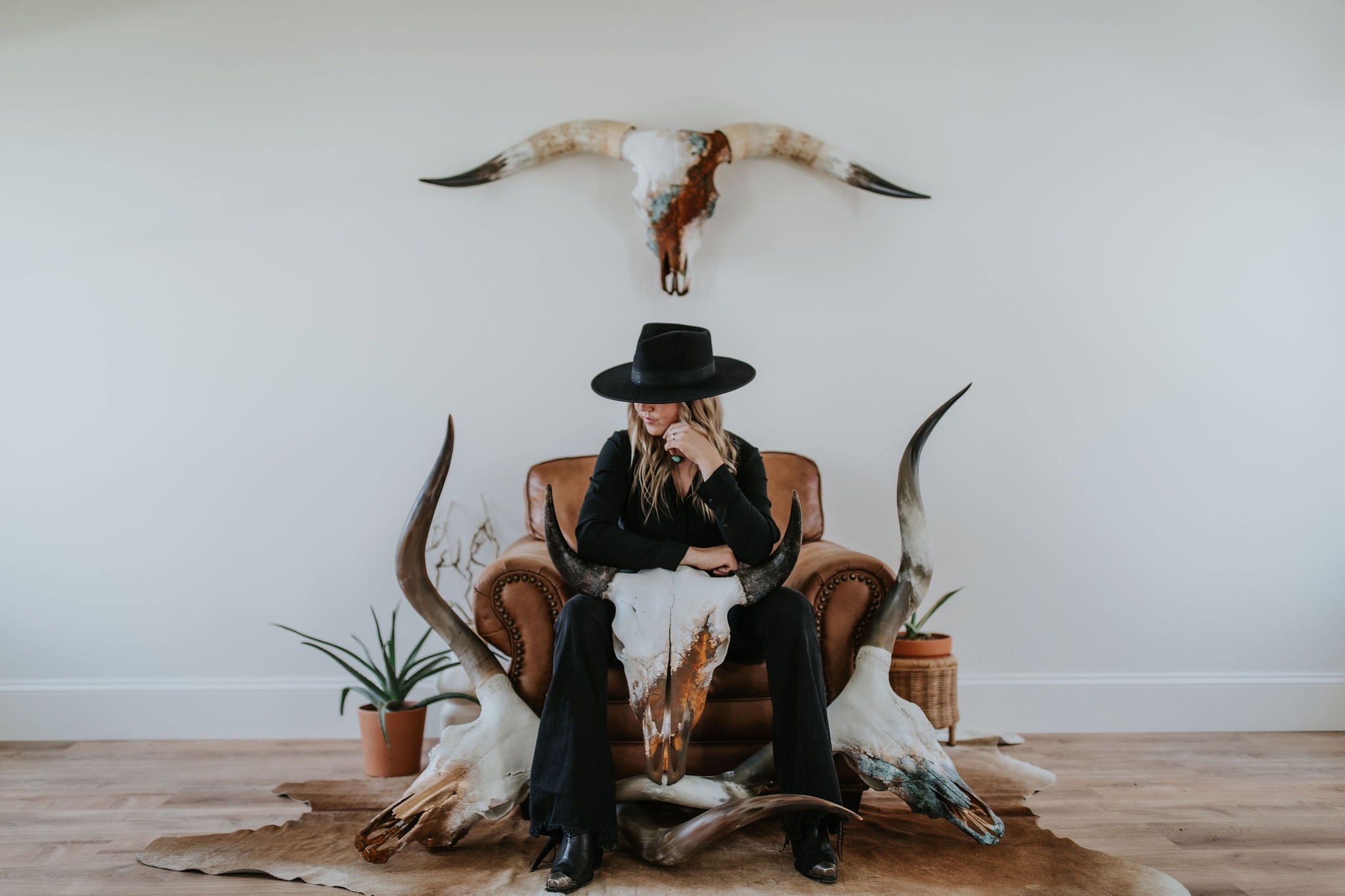 Cassie Everson of Wanderlust Skulls sitting on a chair with two longhorn skulls and a buffalo skull in a room with plants and a wall hung longhorn skull.
